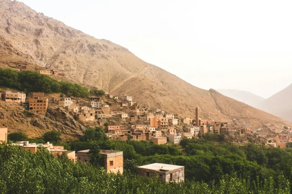 Traditional Berber village houses built into a mountainside in Imlil, with green terraced fields and a mosque minaret in the valley below