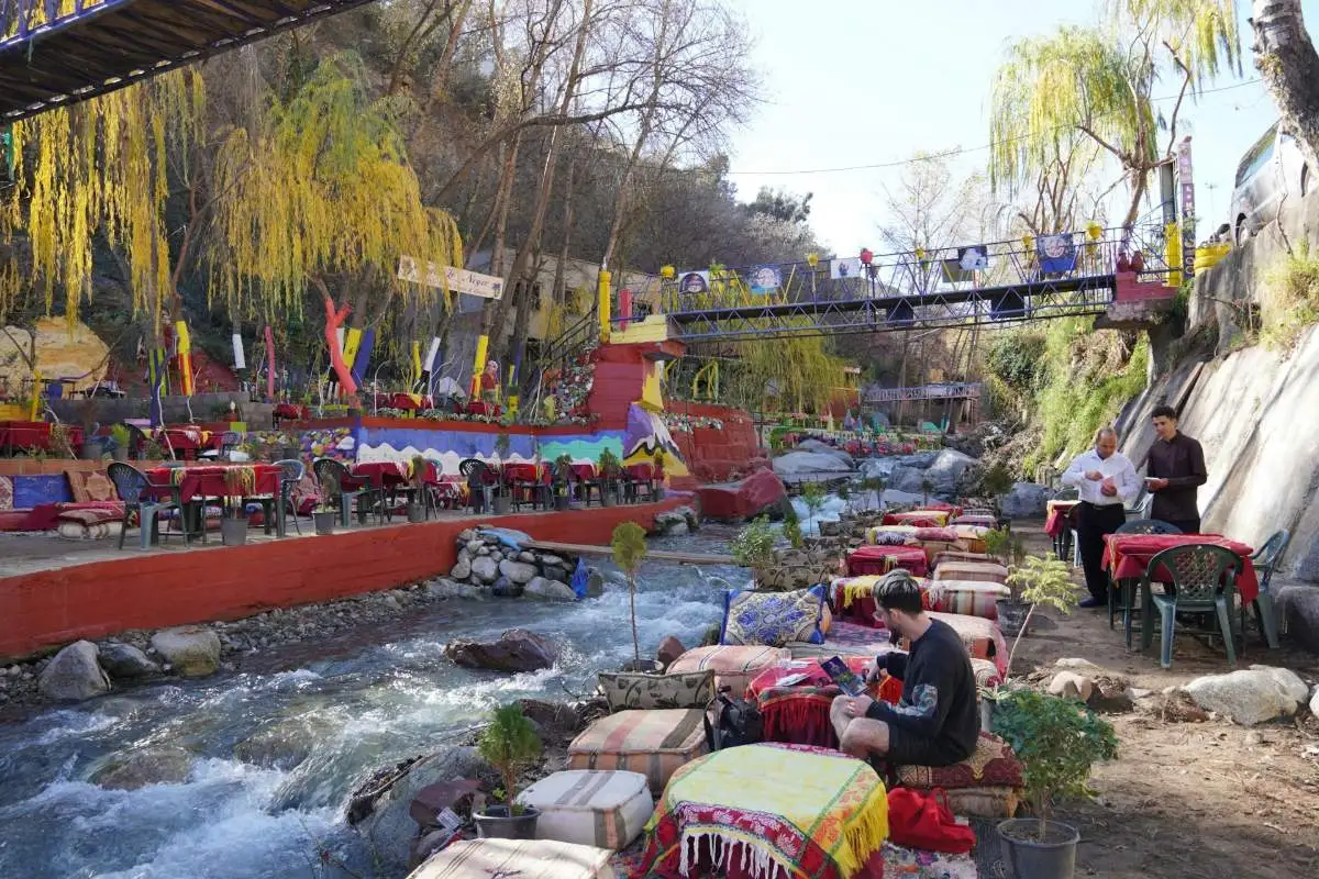 Colorful riverside restaurant seating with cushioned poufs along the Ourika river, with willow trees, a footbridge, and the Atlas Mountains in the background